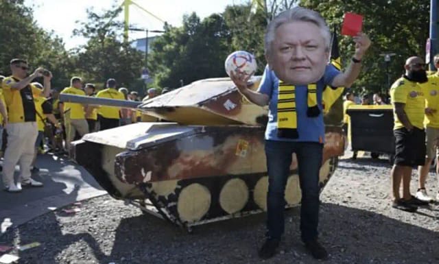 Un tanque ficticio junto a un activista que lleva una máscara con la imagen del director ejecutivo de Rheinmetall, Armin Papperger, y sostiene una tarjeta roja antes de un partido de fútbol en Dortmund, Alemania, el 24 de agosto de 2024. Con el impulso hacia la guerra y el mayor gasto en armas, los directores ejecutivos de armamento como Papperger están esperando que sus ganancias exploten. | Bernd Thissen / dpa vía AP